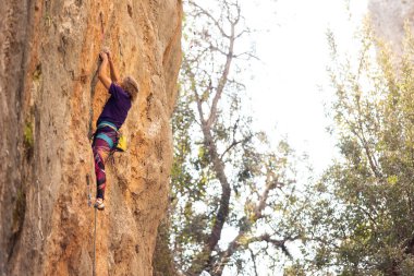Güçlü bir kadın kaya tırmanıcısı uçuruma tırmanır. Kadın, yükseklik korkusunun üstesinden gelerek gücünü ve dayanıklılığını geliştiriyor. Açık hava sporları. Bir kız kayalıklarda antrenman yapıyor.