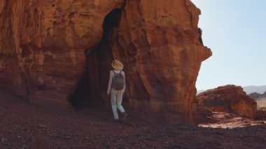 Sırt çantalı bir kız, çölde kırmızı kayaların önünde yürüyor, manzaranın tadını çıkarıyor. Çöl yolculuğu. Timna Ulusal