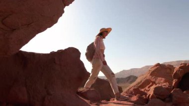 Sırt çantalı bir kız, çölde kırmızı kayaların önünde yürüyor, manzaranın tadını çıkarıyor. Çöl yolculuğu. Timna Ulusal