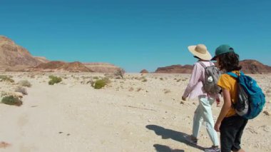 Sırt çantası ve haritası olan bir çocuk çölde annesinin yanında yürüyor. Aile gezisi. Timna Ulusal Parkı. Çocuk.