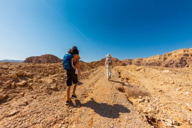 Sırt çantası ve haritası olan bir çocuk çölde annesinin yanında yürüyor. Aile gezisi. Timna Ulusal Parkı. Çocuk aktif olarak zaman geçiriyor. Gezinti ve yürüyüş.