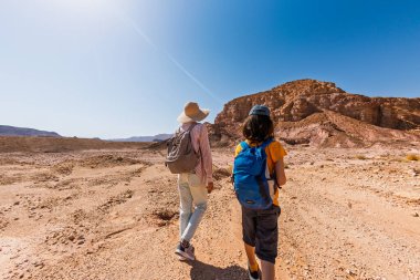 Sırt çantası ve haritası olan bir çocuk çölde annesinin yanında yürüyor. Aile gezisi. Timna Ulusal Parkı. Çocuk aktif olarak zaman geçiriyor. Gezinti ve yürüyüş.