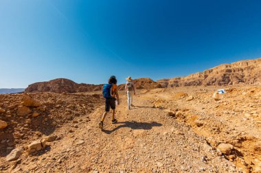 İki aktif gezgin mavi gökyüzüne karşı çölde geziniyor. Anne ve çocuk yürüyüşe çıktı. Yaz kampı. Timna Ulusal Parkı. Çocuk aktif olarak zaman geçiriyor. Gezinti ve yürüyüş.