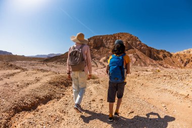 Sırt çantası ve haritası olan bir çocuk çölde annesinin yanında yürüyor. Aile gezisi. Timna Ulusal Parkı. Çocuk aktif olarak zaman geçiriyor. Gezinti ve yürüyüş.