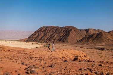 Sırt çantası ve haritası olan bir çocuk çölde annesinin yanında yürüyor. Aile gezisi. Timna Ulusal Parkı. Çocuk aktif olarak zaman geçiriyor. Gezinti ve yürüyüş.