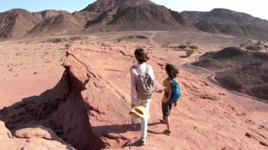 Sırt çantalı bir çocuk dağ yolunda annesinin yanında yürüyor. Aile gezisi. Timna Ulusal Parkı. Çocuk aktif olarak...
