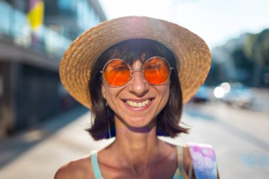 Güneş gözlüklü ve şapkalı neşeli bir kız. Moda kıyafetleri içinde gülümseyen genç bir hippi. Baştan çıkarıcı ve kaygısız kadın şehrin arka planında poz veriyor ve gülümsüyor..