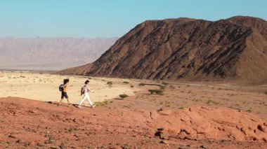 Sırt çantalı bir çocuk dağ yolunda annesinin yanında yürüyor. Aile gezisi. Timna Ulusal Parkı. Çocuk aktif olarak...