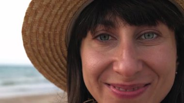 Sahilde duran şapkalı genç bir kadının ön görüntüsü. Gülümsüyor ve kameraya bakıyor. Mutlu kız.