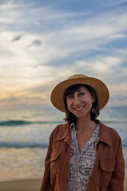 Sahilde duran şapkalı genç bir kadının ön görüntüsü. Gülümsüyor ve kameraya bakıyor. Mutlu kız gülümsüyor. Rahatlama ve macera.