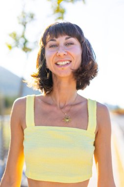 Havalı yaz kıyafetleri içinde, yüzü piercingli, gülümseyen genç bir esmer. Güneşli bir günde dışarıda poz veren baştan çıkarıcı, kaygısız kadın. Açık havada pozitif model. Yaz havası.