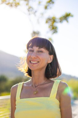 Zarif sarı elbiseli, güzel, gülümseyen esmer kız. Günbatımında dışarıda poz veren baştan çıkarıcı, kaygısız kadın. Güneşli bir günde pozitif bir model. Yaz havası.