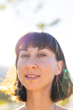 Havalı yaz kıyafetleri içinde, yüzü piercingli, gülümseyen genç bir esmer. Güneşli bir günde dışarıda poz veren baştan çıkarıcı, kaygısız kadın. Açık havada pozitif model. Yaz havası.