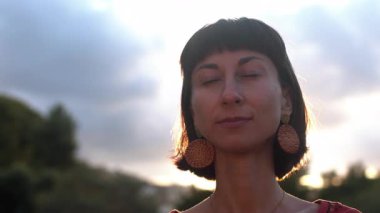 Kısa saçlı, doğal güzellikte bir kadının yakın plan portresi. Bir yaz parkında gözleri kapalı duruyor ve eğleniyor.