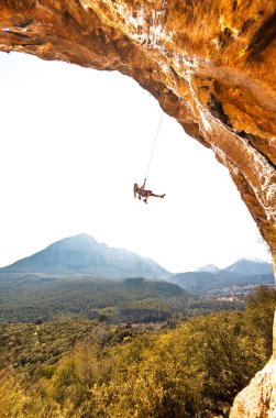 Dişi bir kaya tırmanıcısı. Bir kaya tırmanışçısı bir ipe asılır gökyüzünün arka planına ve güzel dağlara karşı. Bir kız tırmanma rotasında düşer. Kaya tırmanışı ve dağcılık. Açık hava sporları. Mağara.