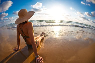 Şapkalı ve mayolu bir kız deniz kıyısındaki kumda oturur ve dalgaların ve günbatımının tadını çıkarır. Arkadan bak. Tatil ve yaz havası. Yaz tatili kavramı.