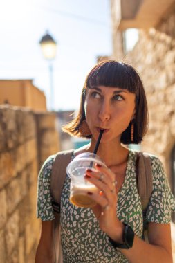 Güneşli bir yaz gününde şehir sokaklarında dikilirken pipetle kokteyl içen mutlu genç bir kadının portresi. Neşeli bir bayan şehir caddesinde soğuk bir şeyler içmekten hoşlanır. Paket kahve..