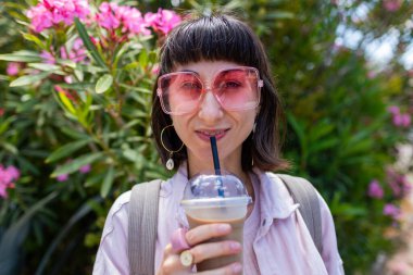 A smiling, happy young woman enjoys a drink outdoors. She's relaxing, taking a break. A girl drinks coffee while walking along a city street. City life on the street.