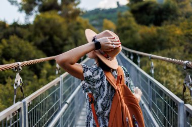 Bir kadın turist nehrin üzerindeki köprüden geçiyor. Genç bir kadın, sonbahar ormanının önündeki bir nehrin karşısındaki asma köprü boyunca yürüyor. Şapkalı kız..