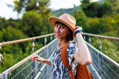 Sırt çantalı bir kız. Şapkalı ve sırt çantalı genç bir kadın nehrin üzerindeki asma köprü boyunca yürüyor. Seyahat ve macera.