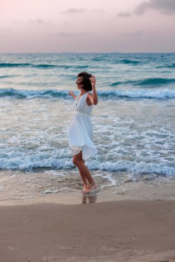Beyaz yazlık elbiseli mutlu genç kız gün batımında sahilde yürüyor. Kumsalda dans eden kız..