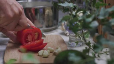 Parlak bir mutfak ortamında, tahta kesme tahtasının üzerinde kırmızı bir domatesi bıçakla dilimleyen bir adam. Adam yemek yapıyor. Güzel mutfak..