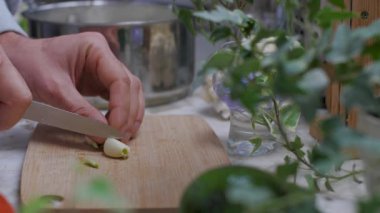 Bir adam sarımsağı tahta bir kesme tahtasının üzerinde bıçakla doğruyor. Sahne, sıcak bir mutfakta geçiyor. Sıcak bir atmosfer yaratıyor.