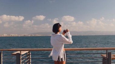 Elinde kamera olan bir kız denizin kenarında durup fotoğraf çekiyor. Şık bir kadın fotoğraf makinesiyle fotoğraf çekiyor. Kadın bir blogcu.