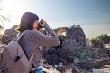 Kameralı bir kız. Blogu ve sosyal medyası için sırt çantalı bir turist. Bir turist fotoğrafları antik taş kalıntılarını gösteriyor..