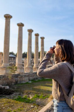Kameralı bir kız. Blogu ve sosyal medyası için sırt çantalı bir turist. Bir turist fotoğrafları antik taş kalıntılarını gösteriyor..