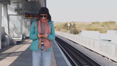 Genç bir kadın telefonunu kullanarak platformda duruyor. Yolcunun biri telefonundaki uygulamayı kullanarak tren tarifesini kontrol ediyor. Bir kız...
