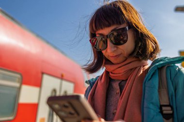 Genç bir kadın telefonunu kullanarak platformda duruyor. Yolcunun biri telefonundaki uygulamayı kullanarak tren tarifesini kontrol ediyor. Karakolda bir kız var..