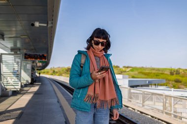 Platformda elinde telefonla tren bekleyen genç bir kadın var. Yolcunun biri telefonundaki uygulamayı kullanarak tren tarifesini kontrol ediyor. Karakolda bir kız var..