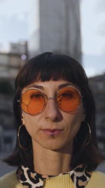 Dikey çekim. Yakın plan. Yuvarlak güneş gözlüklü genç bir kadın kameraya gülüyor ve gülümsüyor. Şık bir kızın portresi..