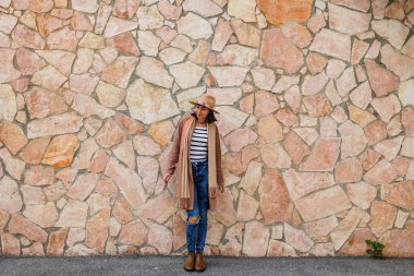 Güzel, gülümseyen bir modelin portresi. Bir şehir duvarının arkasında şapka takıyor ve şık kıyafetler giyiyor. Bir kadın sokakta bir duvarın yanında poz veriyor. Komik ve pozitif kadın dışarıda eğleniyor..