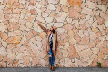 Güzel, gülümseyen bir modelin portresi. Bir şehir duvarının arkasında şapka takıyor ve şık kıyafetler giyiyor. Bir kadın sokakta bir duvarın yanında poz veriyor. Komik ve pozitif kadın dışarıda eğleniyor..