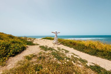 Yaz havası. Kollarını kaldırmış, beyaz elbiseli mutlu genç bir kadın deniz kıyısında yürüyor ve denize bakıyor. Kumsalda dinlenmek..