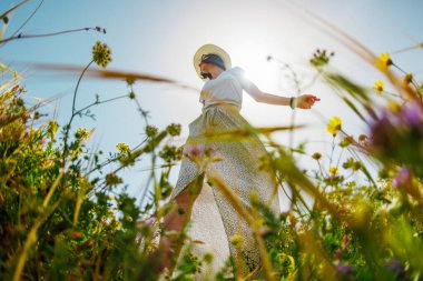 Yaz havası. Mavi gökyüzü ve çiçeklerle dolu bir arka planda poz veren genç ve neşeli bir kız. Güzel bir kız çiçekler arasında dans eder. Çiçeklerin arasında mutlu bir kız, açık havada. bahar modası.