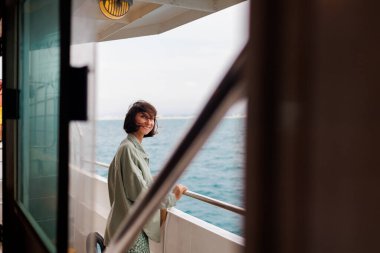 Genç bir kadın bir yolcu gemisinin güvertesinde duruyor ve batan güneşe bakıyor. Genç ve güzel bir esmer, güzel bir mavi denizin arka planında bir yatla seyahat ediyor. seyahat ve macera.