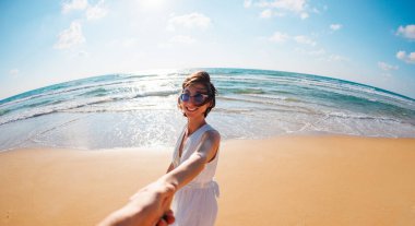 Birbirlerine aşık çiftler sahilde eğleniyorlar. Beni takip edin. Genç ve mutlu bir kadın erkeklerin elini çekiyor. Elbise giymiş güzel bir kız sahilde yürüyor, bir erkeğin elini tutuyor, romantik bir yürüyüş. Sevgililer Günü.