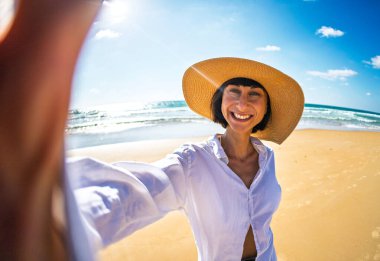 Neşeli genç bir kadın, tropik bir plajda tatildeyken akıllı telefonuyla mutlu bir selfie çekiyor. Şapkalı mutlu bir manken tatilinin tadını çıkarır. Yaz yaşam tarzı.