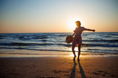 Sahilde dans eden kız. Çekici, mutlu, elbiseli ve şapkalı genç bir kadın gün batımında boş vakitlerin tadını çıkarıyor. Bahar yaşam tarzı kavramı, özgürlük.