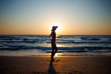 Gün batımında sahilde yürüyüş yapan bir kız. Gün batımına bakan bir kadının silueti. Yaz havası. Sahildeki kız..