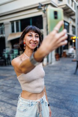 Sıradan giysiler içinde bir kızın resmi, gülümsüyor, şehir caddesinde yürüyor, tatillerde seyahat ediyor ve selfie çekiyor. Kız selfie çekiyor..