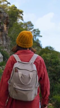 Dikey video. Bir kız gülümsüyor. Mutlu bir turist yürüyüşü. Dişi bir turist sırt çantasıyla bir dağ zeminine doğru yürüyor.