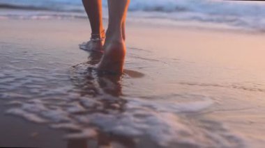 Bir kız plaj boyunca yürür. Sahilde çıplak ayaklı bir kadının yakın çekimi. Kadın su boyunca yürüyor ve eğleniyor.