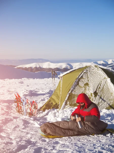 Un hombre se sienta en un saco de dormir cerca de la tienda y raquetas de nieve . — Foto de Stock