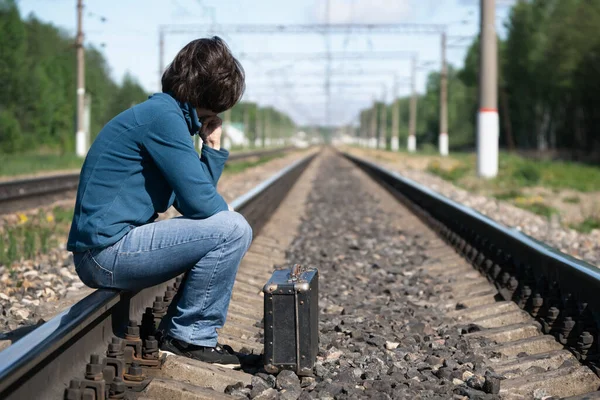 Mujer con maleta vieja se sienta en un carril de ferrocarril y espera un tren tarde en la mañana de verano — Foto de Stock