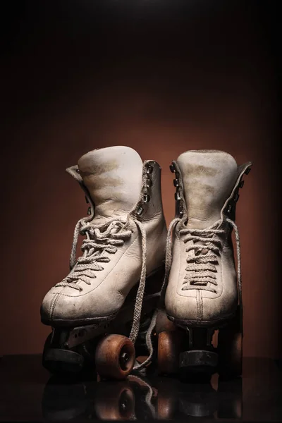 Old heavily used worn-out pair of white roller skates showing their boot tongues with some dirt and untied shoelace on reflective surface and warm brown background with upper space for titles