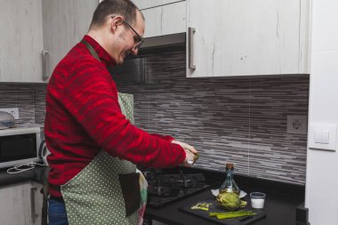 Kırmızı postlu ve yeşil önlüklü bir adam mutfakta enginar ve kuşkonmaz pişiriyor. Enginar kesiyor..